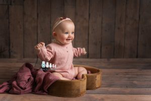 professional baby portrait of baby sitting in a heart bowl on a wooden backdrop smiling off camera wearing a pink sleeved outfit and headband. By edinburgh baby photographer Beautiful Bairns Photography