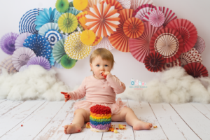 cake smash photo of little girl celebrating 1st birthday with a rainbow themed cake smash with rainbow cake and rainbow paper fans behind her by cake smash photographer beautiful bairns photography edinburgh