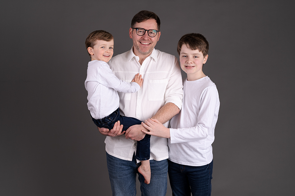 professional family portrait od dad and two sons. All wearing white shirts and denim jeans in front of a grey backdrop. Photograph by Beautiful Bairns photography Edinburgh family photographer