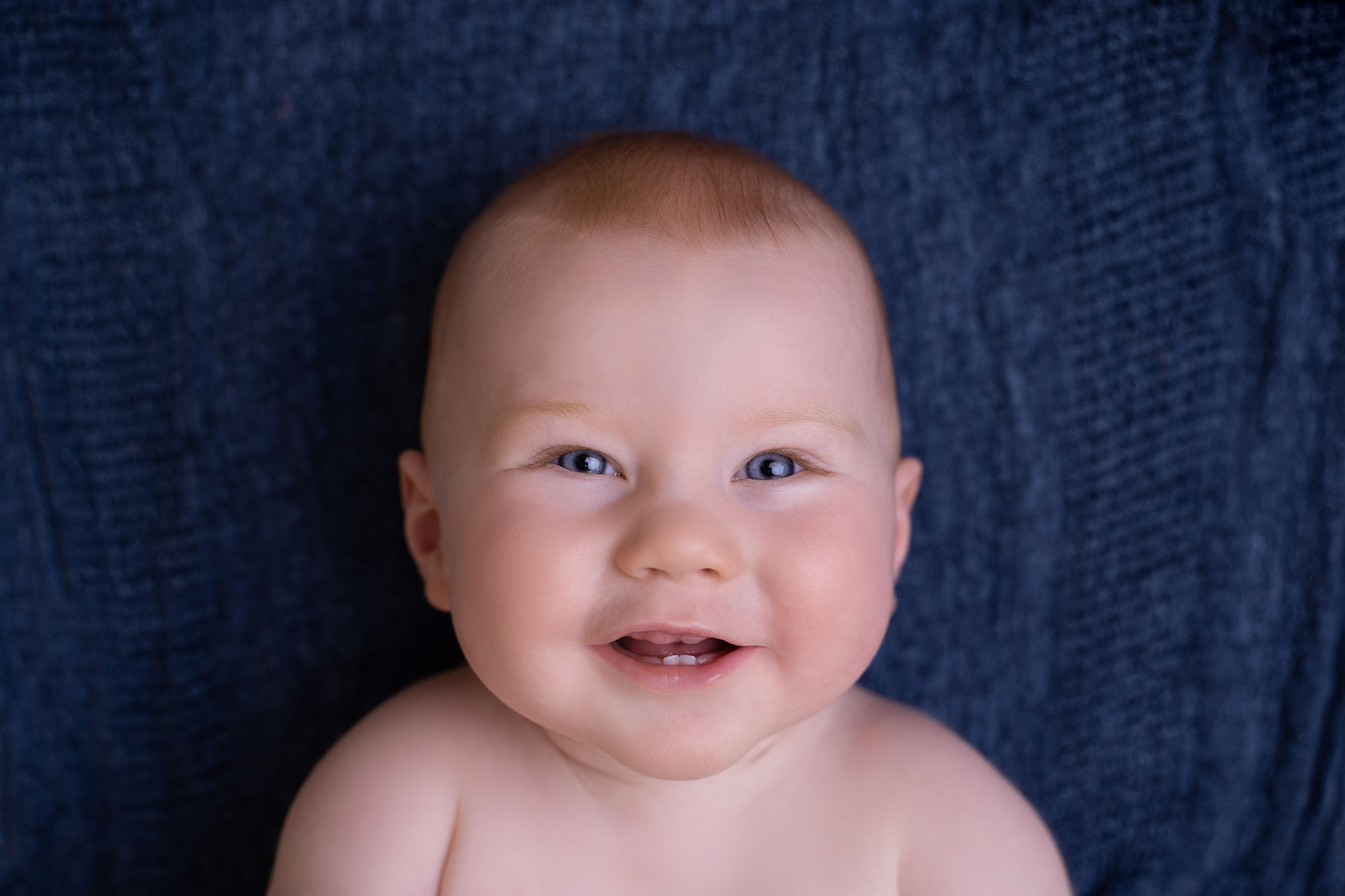professional baby portrait of young baby laying on back smiling at the camera laid on a blue fabric